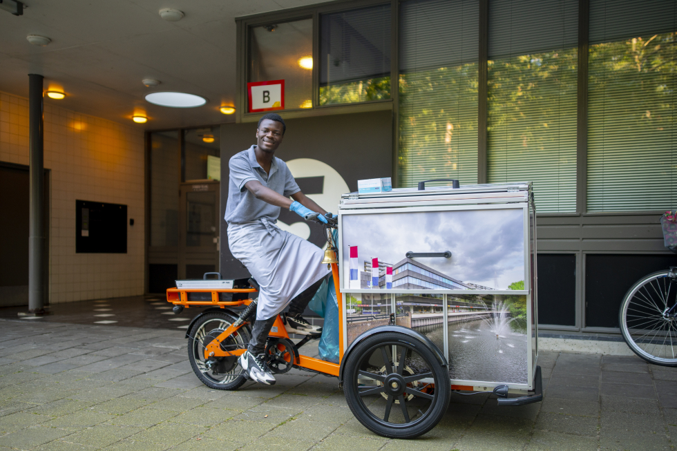 Een vrijwilliger die op de fiets van de soepkar zit. Op de soepkar staan drie foto's van de wijk Vollenhove. De soepkar staat voor de portiek van een flat. 