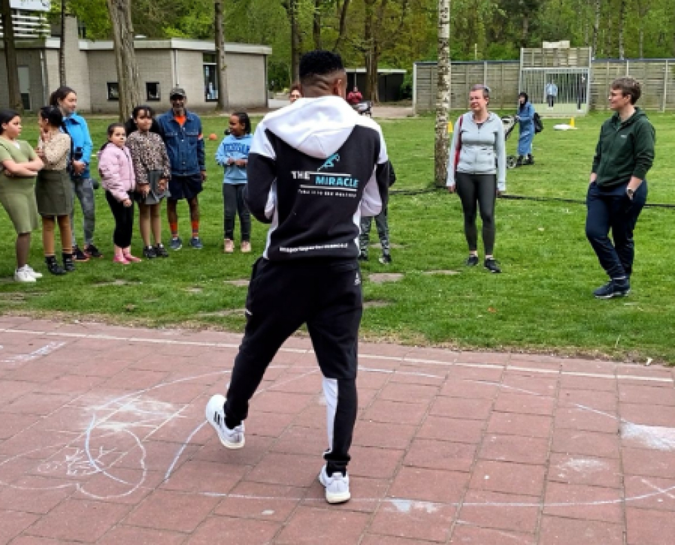 Gennaro staat in een park voor een groep mensen die meedoen aan bootcamp. Hij heeft sportkleding aan met de bedrijfsnaam The Miracle Sports Performance