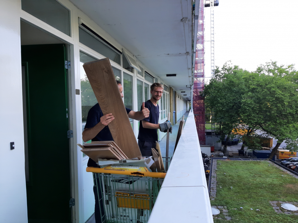 Actieve huurder Laurens aan het werk in de L-flat. Hij staat samen met iemand op de gallerij van de L-flat en ze hebben een boodschappenkarretje met veel houten planken.