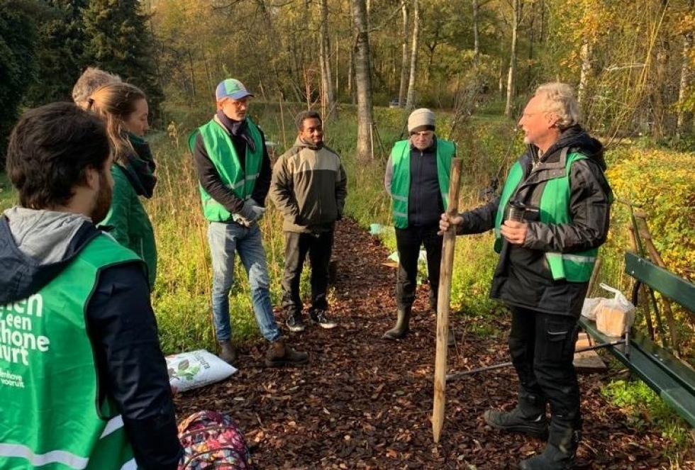 Zes vrijwilligers staan in de Voedselbosrand en krijgen uitleg wat zij kunnen planten. Ze hebben allemaal groene hesjes aan.