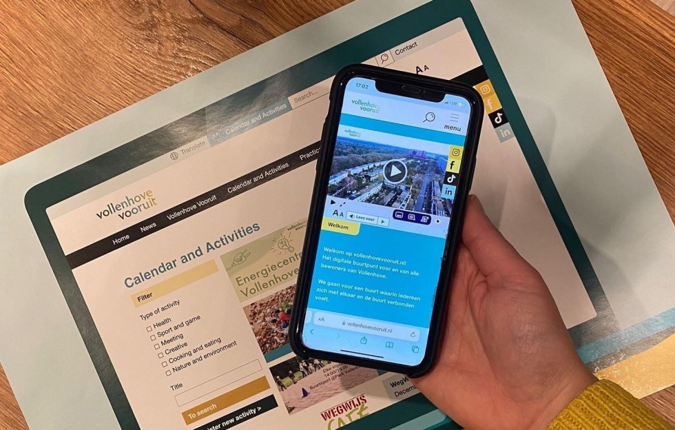 Op de achtergrond is een print te zien van de homepage van de Engelse versie van de Agenda en Activiteiten-pagina van het digitaal buurtpunt. Op de voorgrond is een hand te zien die een telefoon vastheeft met de homepage van het digitaal buurtpunt. 