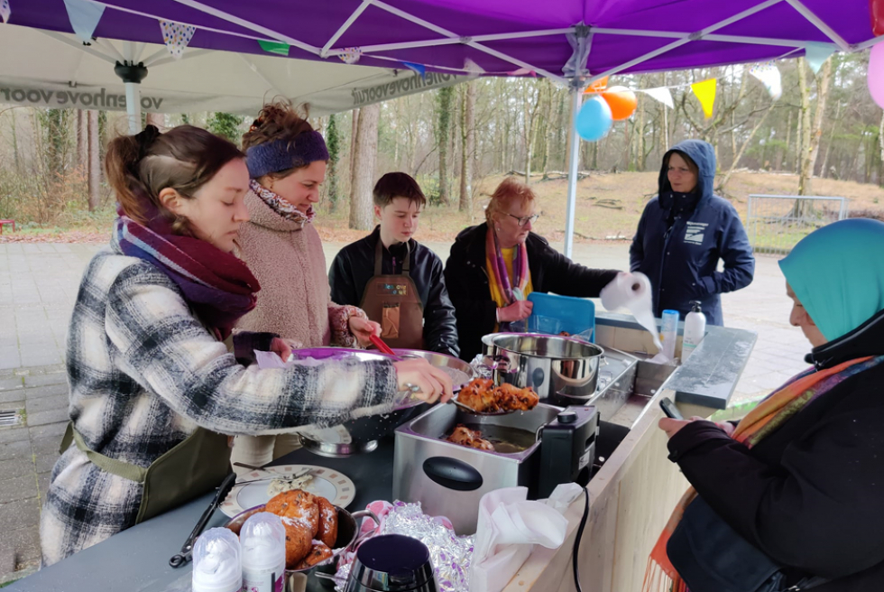 Een aantal bewoners die voor de oliebollenkraam de oliebollen bakken met een frituurpan. Ze staan onder een paarse partytent met vlaggetjes