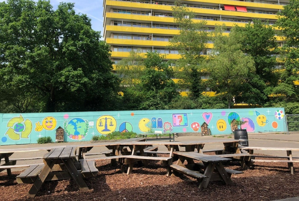Het ontmoetingsplein met de buurtboomtafel en de graffiti/versierde muur en gele Geroflat op de achtergrond.