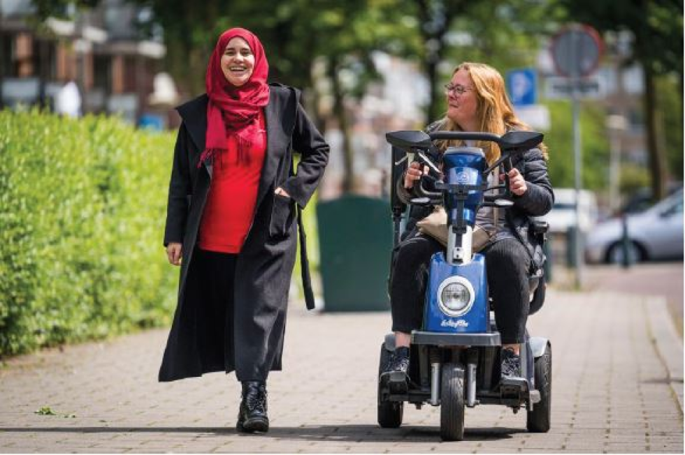 Foto van een vrouw in een scootmobiel en iemand in opleiding die haar helpt