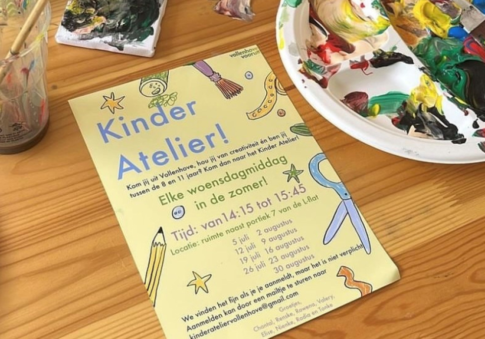 Foto van een flyer voor het Kinder Atelier op een tafel met kwasten en verf. Op de flyer staat praktische informatie over waar en wanneer het Kinderatelier is.