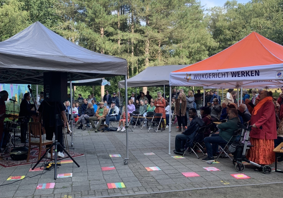 Een foto van het wereldmuziekfestival met links de band en rechts publiek op stoeltjes op 't Ontmoetingsplein.