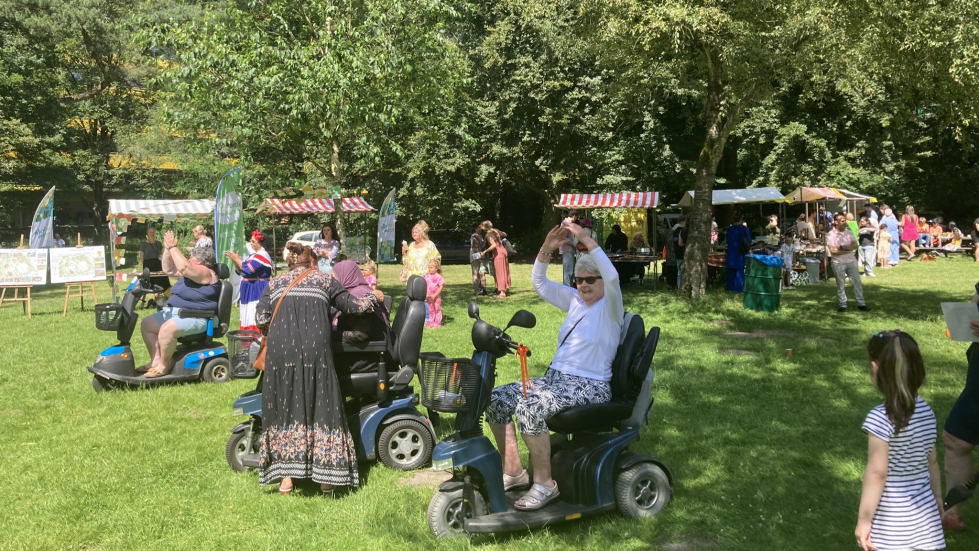 Een foto van het buurtfeest in het park in 2024. In de voorgrond rijden meerdere bezoekers op scootmobielen over het gras. In de achtergrond staan kraampjes, vlaggen en groepjes mensen die deelnemen aan of kijken naar de activiteiten.